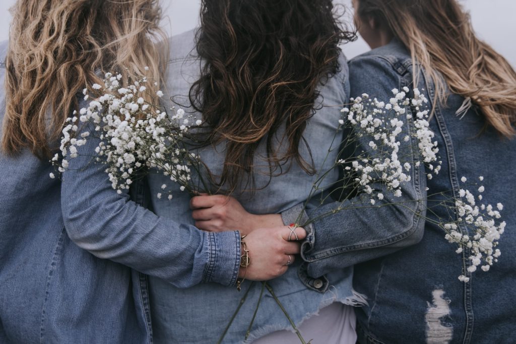three women facing the opposite direction wearing denim jackets and holding white flowers