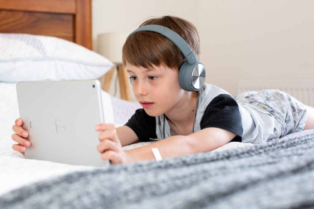 young boy laying on bed wearing headphones and watching on his tablet device