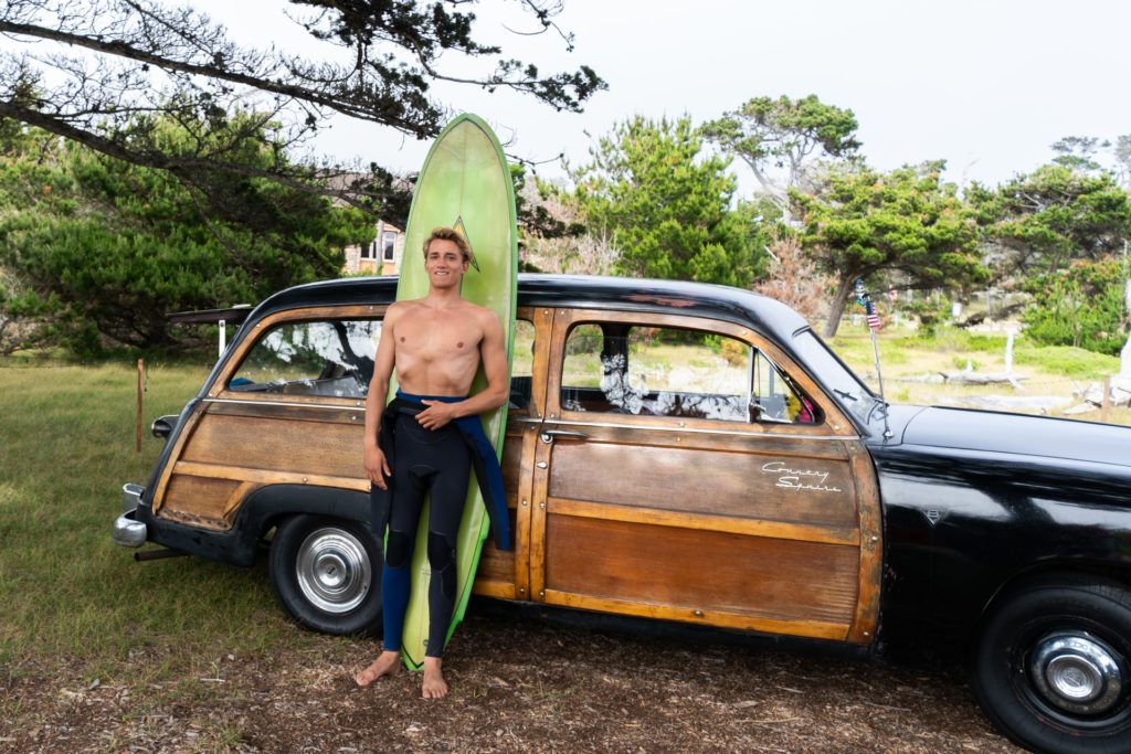 male surfer in wetsuit standing in front of his surfboard leaning against vintage car
