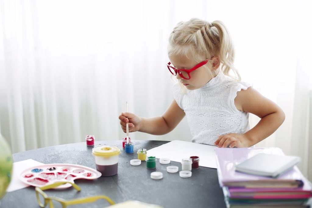 young girl wearing red glasses doing artwork and painting on white paper with poster paint