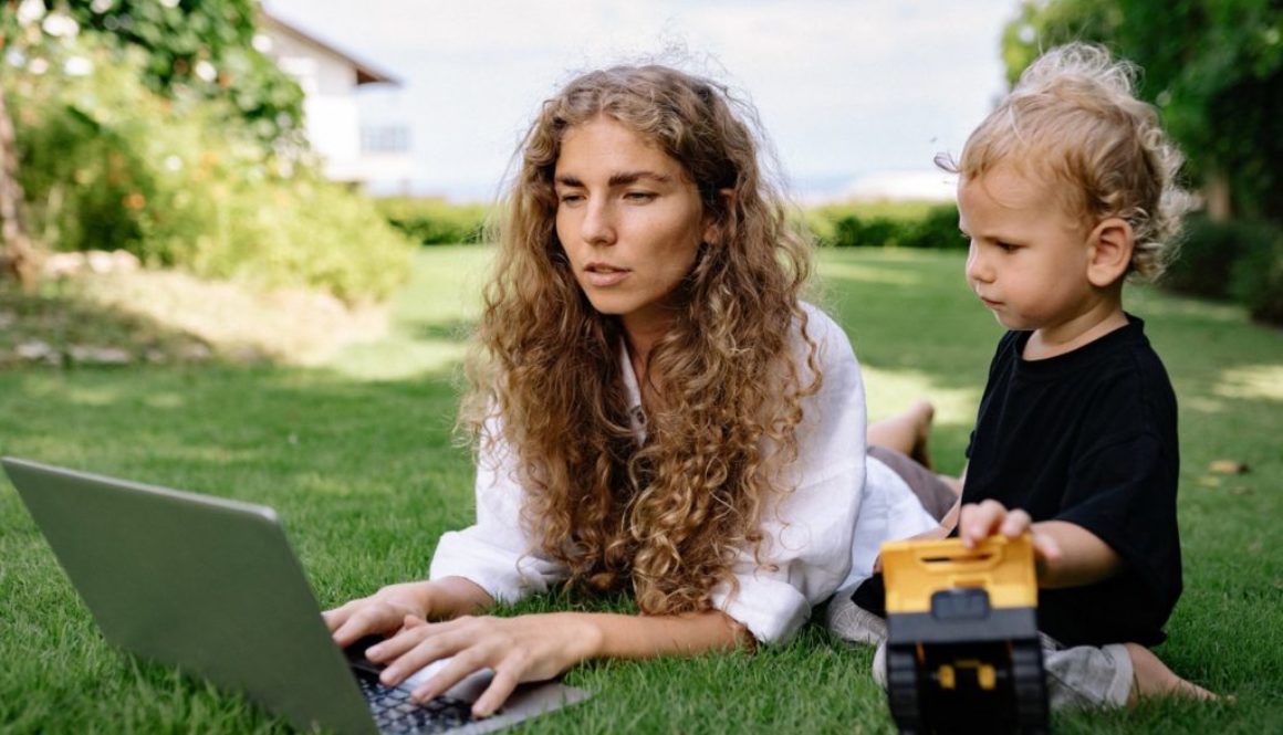 working mom on her laptop lying on the grass with young son playing with toy truck
