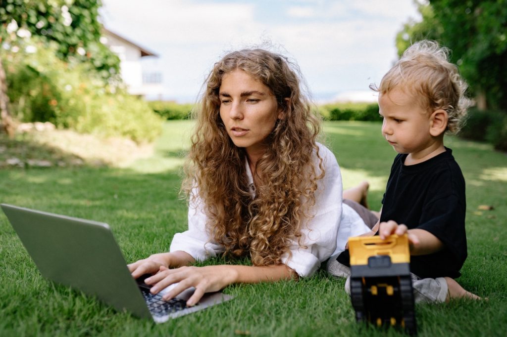 working mom on her laptop lying on the grass with young son playing with toy truck