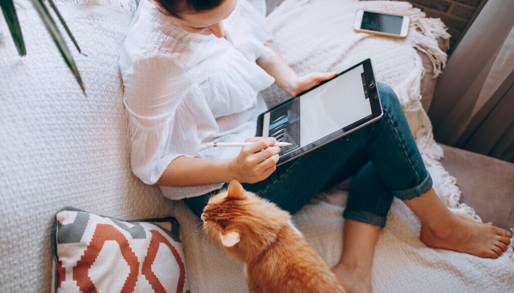 A woman working on her couch on a tablet with a cat next to her