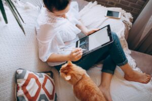 A woman working on her couch on a tablet with a cat next to her